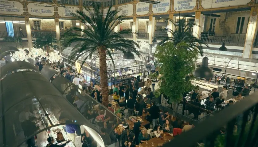 Un bullicioso mercado interior con palmeras, gente comiendo y comprando. El ambiente recuerda a un mercado de la imprenta, lleno de vida y actividad.