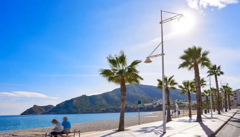 Pareja sentada en un banco en la playa cap blanc, con palmeras y montañas al fondo bajo un cielo azul brillante.