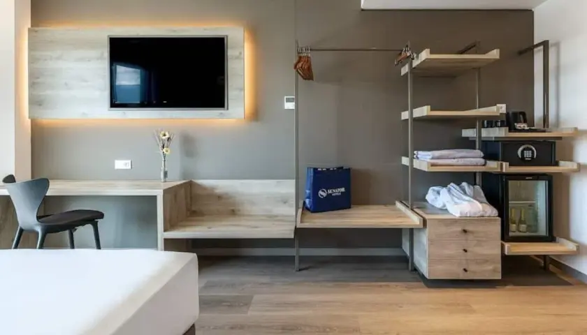 Habitación moderna con TV, escritorio, armario abierto con minibar y caja fuerte. Una bolsa azul con el logo de Senator Hotels descansa sobre un banco.