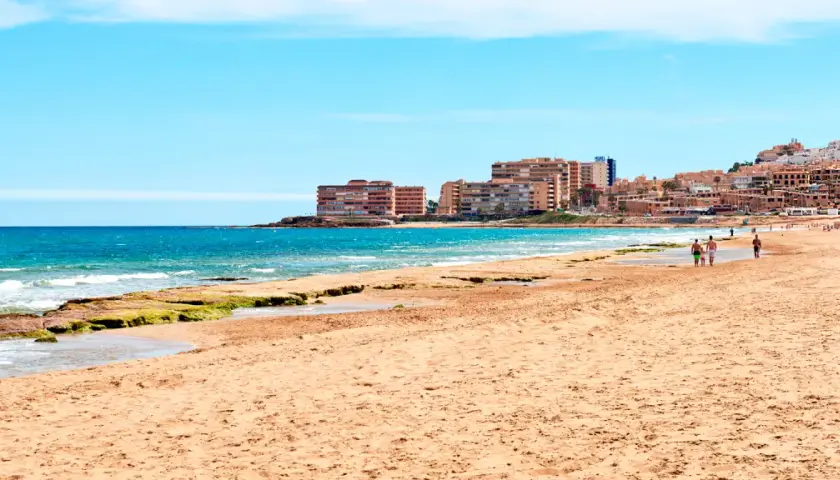 Gente pasea por la **playa de la Mata** con edificios residenciales al fondo y el mar azul.