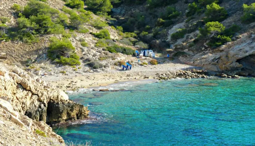 Agua turquesa en la cala Almadrava de Benidorm, con rocas y vegetación en la ladera. Hay algunas sillas y sombrillas en la playa.