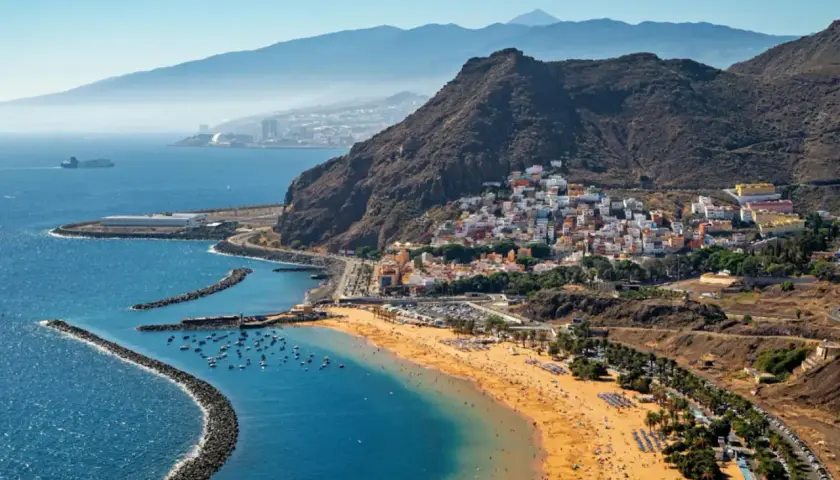 Vista aérea de una playa dorada y un pueblo costero con casas blancas y coloridas en la costa blanca España. Montañas y el mar azul completan la escena.
