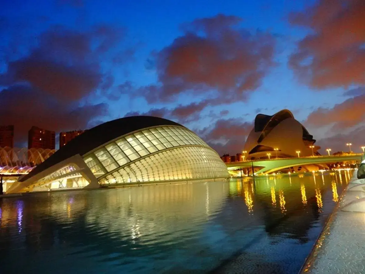 La **Ciudad de las Artes y las Ciencias** de Valencia, con sus icónicos edificios futuristas, se refleja en el agua al anochecer, bajo un cielo dramático.