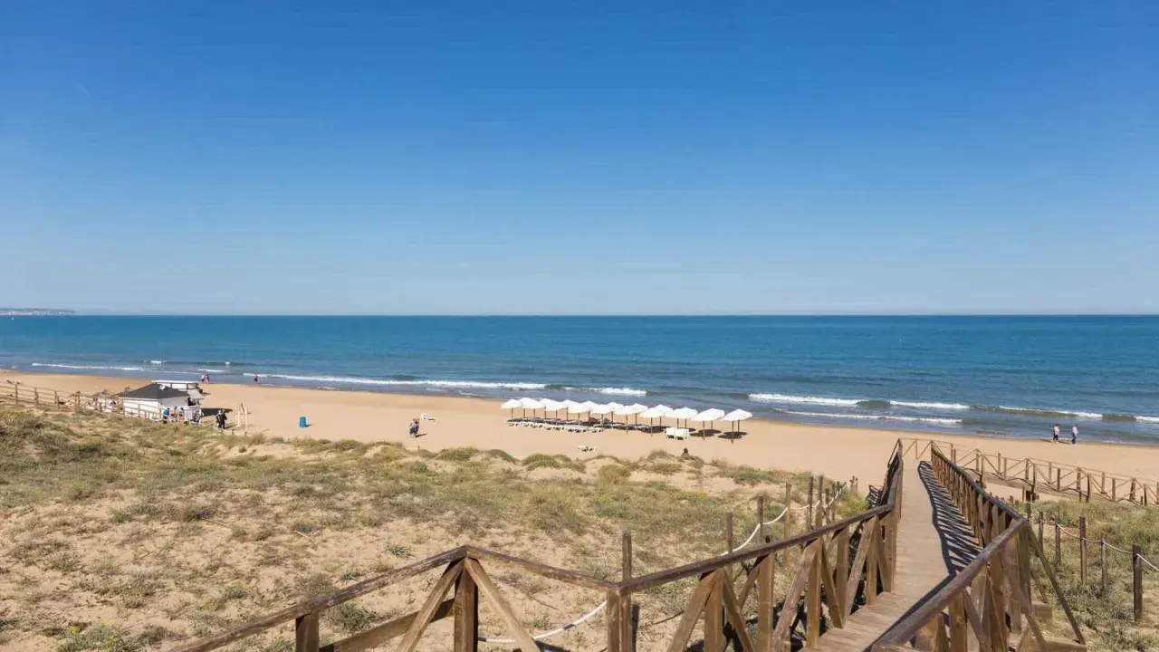 Pasarela de madera hacia la playa de Miramar, con sombrillas blancas alineadas y el mar azul bajo un cielo despejado.