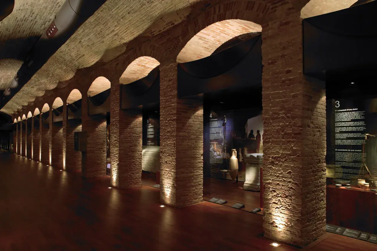 Pasillo del museo de historia de Valencia, con arcos de ladrillo, iluminación tenue y vitrinas con artefactos antiguos.