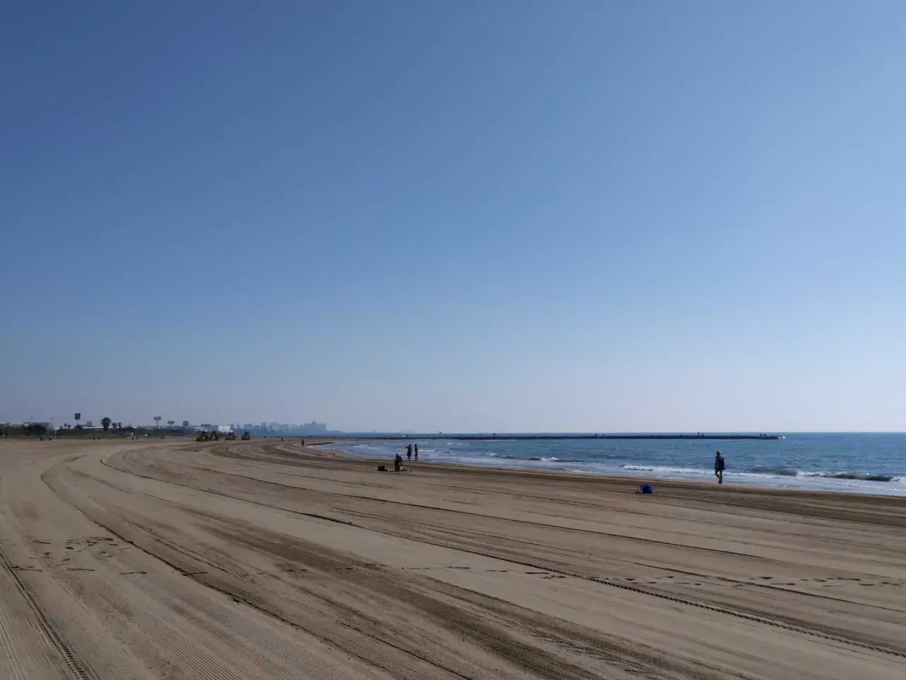 Amplia y tranquila playa de Meliana con arena marcada por vehículos. El mar azul se extiende hasta el horizonte, con algunas personas paseando.