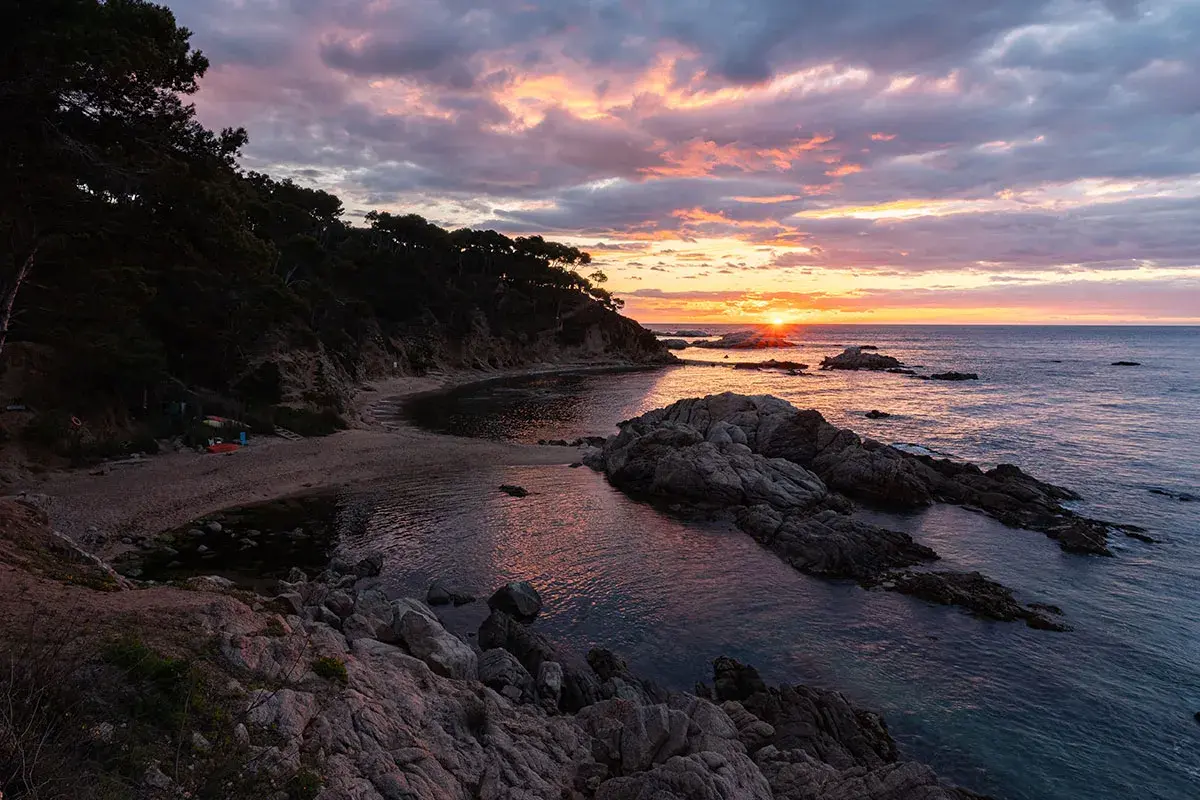 Atardecer en la punta estrella Moraira, con el sol asomando entre rocas y el mar reflejando los tonos rosados del cielo.