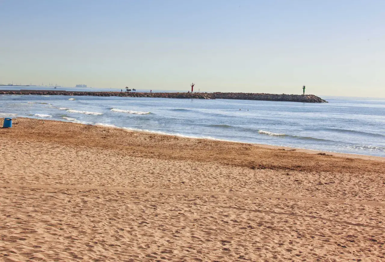 Arena dorada en la playa Port Saplaya, con el mar azul y un espigón con faros.