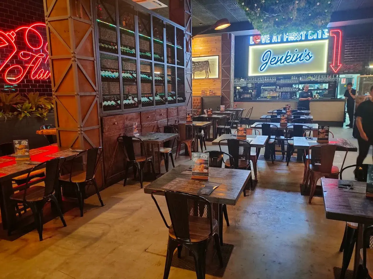 Interior de Jenkins Valencia, un restaurante con mesas de madera, sillas metálicas y un letrero de neón que dice "Love at First Bite Jenkins".