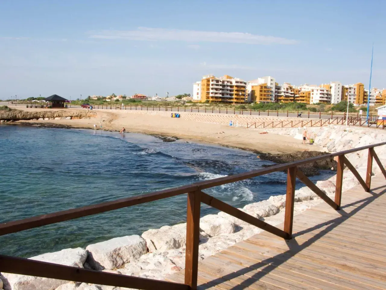 Paseo de madera junto al mar en la cala Piteras. Edificios modernos y playa de arena dorada.