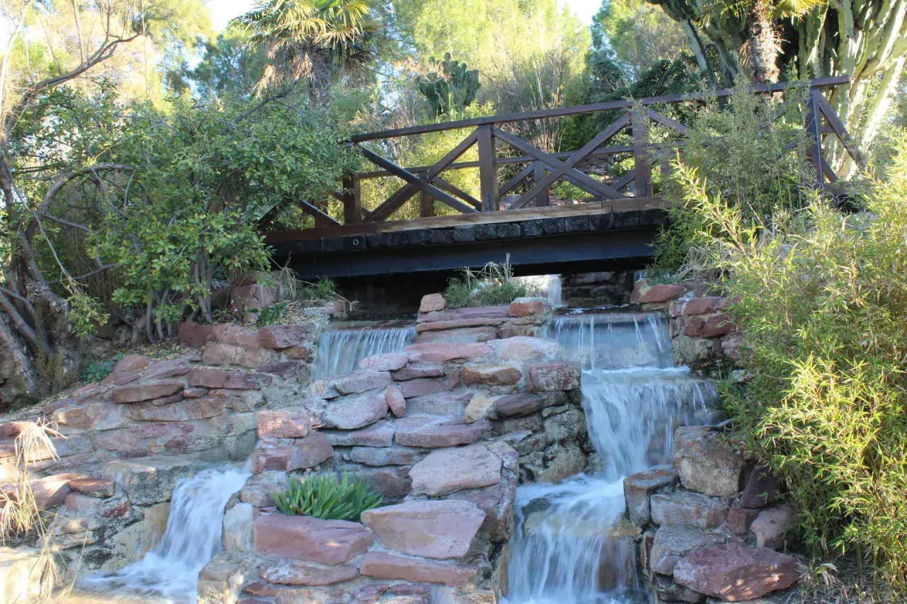 Puente de madera sobre cascadas en el parque de Benicalap, rodeado de vegetación exuberante y rocas rojizas.