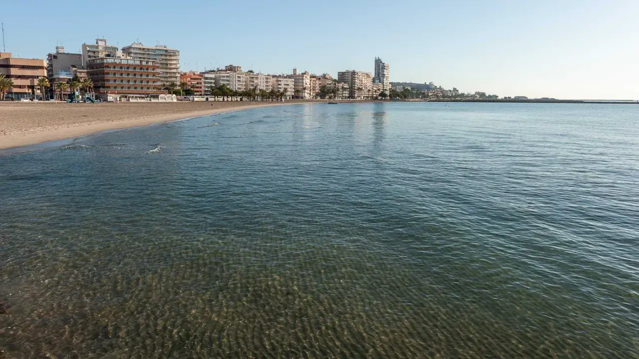 El mar Mediterráneo baña la arena dorada de la playa Levante en Santa Pola, con edificios modernos y palmeras al fondo.