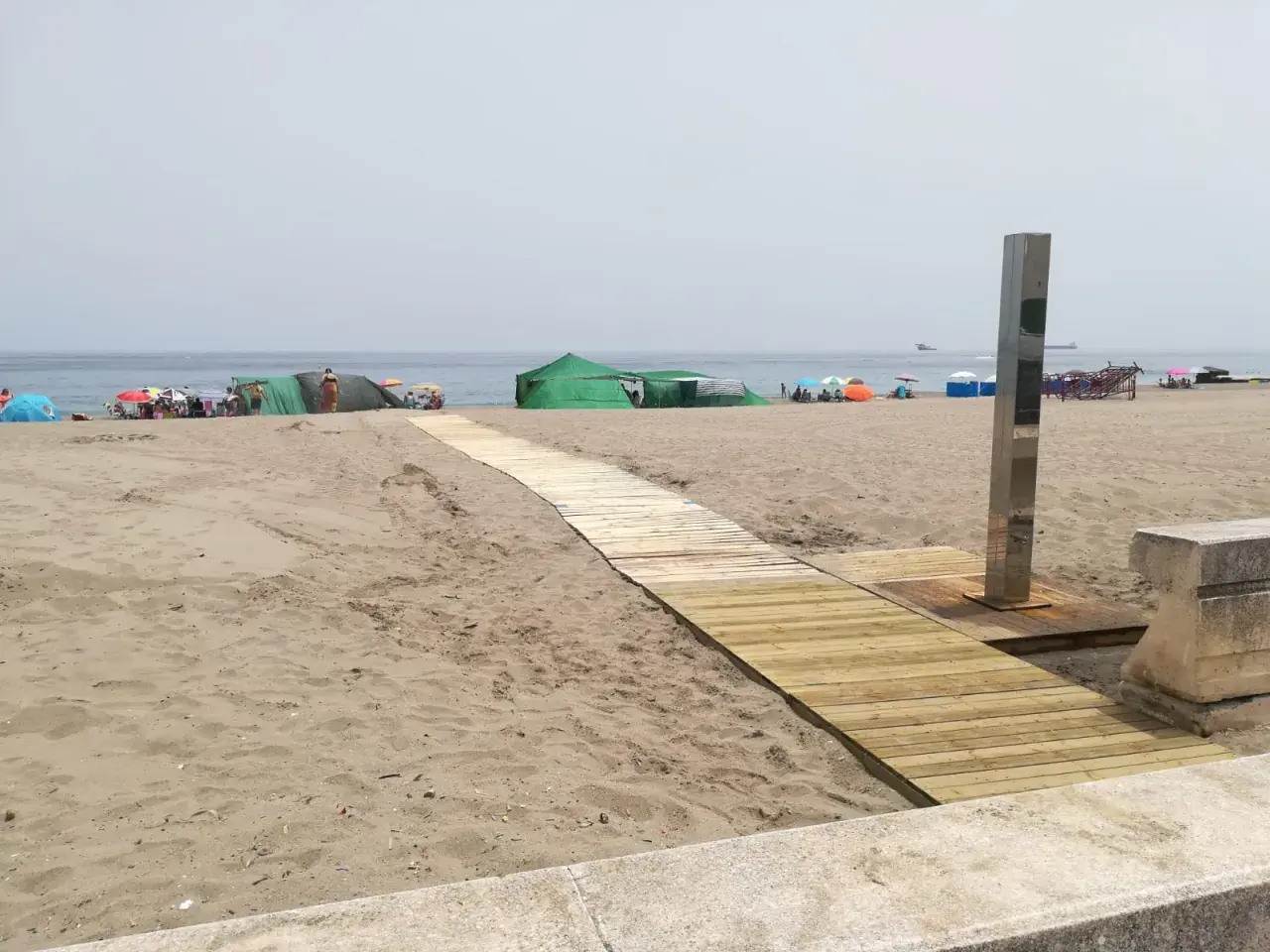 Pasarela de madera hacia las playas con duchas en Alicante. Gente disfrutando del sol bajo sombrillas y tiendas verdes.
