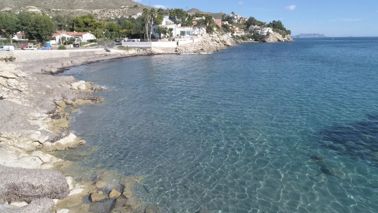 Aguas cristalinas bañan la costa rocosa de cala Piteres, con casas blancas y palmeras en la ladera.