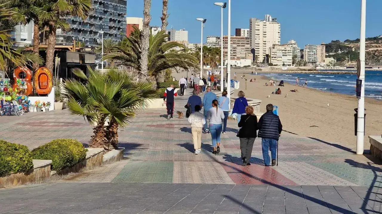 Gente paseando por el **paseo marítimo de Calpe**, con palmeras, edificios altos y la playa al fondo.