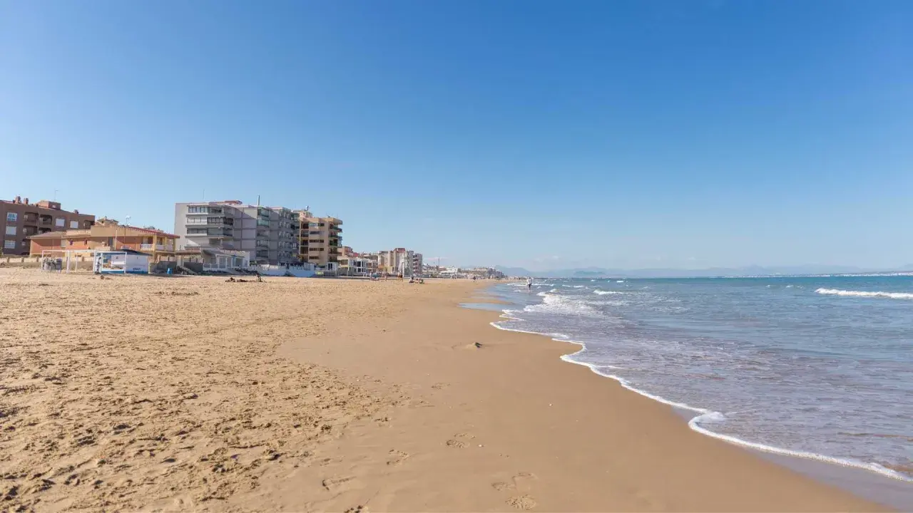 Arena dorada y mar azul en la playa La Roqueta, con edificios de apartamentos y montañas al fondo.