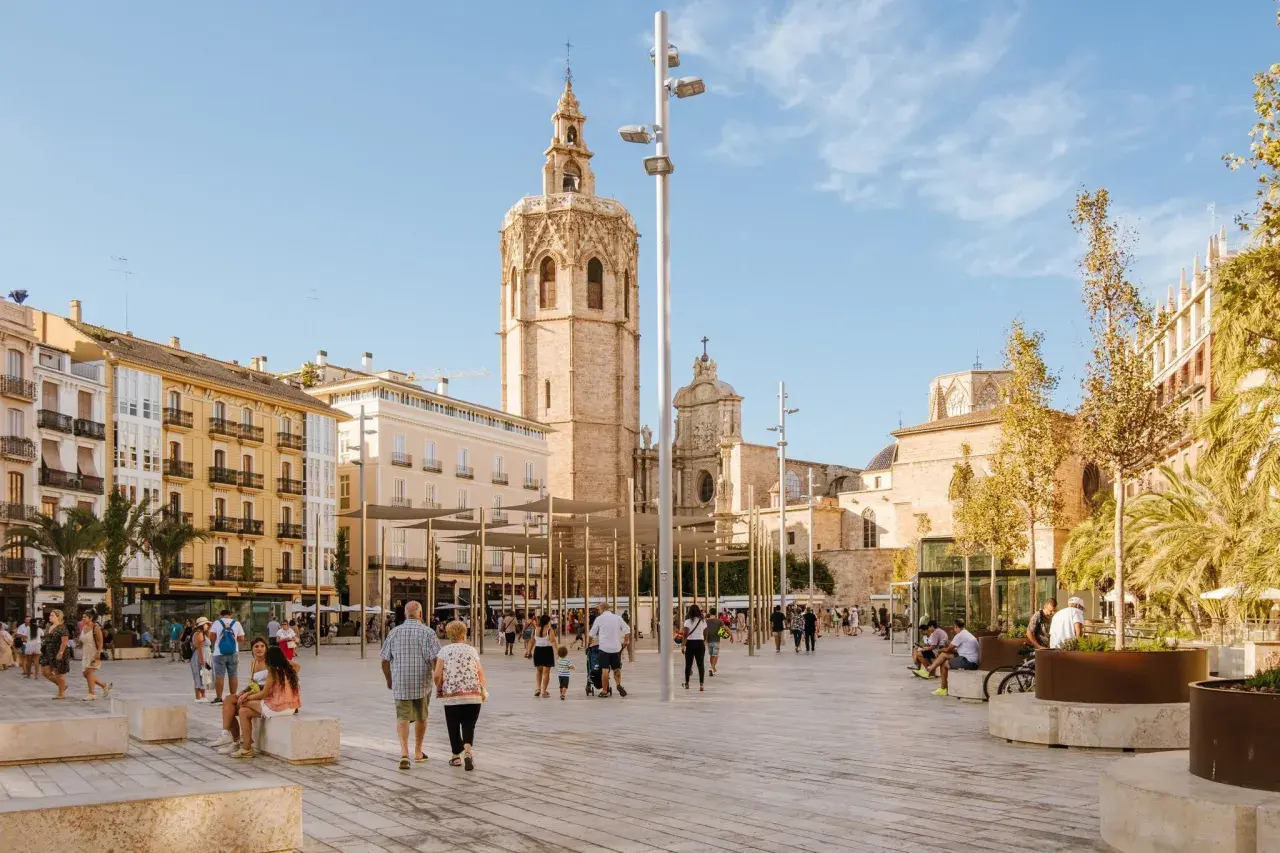 Gente pasea por la plaza frente a la Catedral de Valencia, con el icónico campanario del Miguelete dominando el cielo azul.