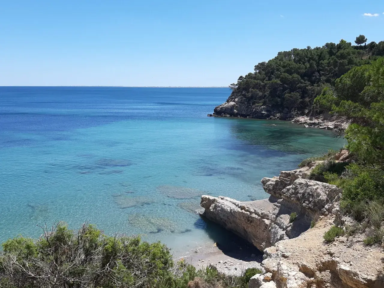Aguas cristalinas y rocas en la playa del Perelló, con vegetación verde y un cielo azul despejado.