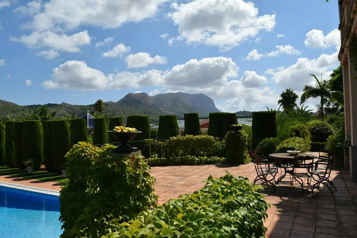 El jardín de l'albarda, con piscina azul, setos verdes, mesa y sillas, y montañas al fondo bajo un cielo nublado.