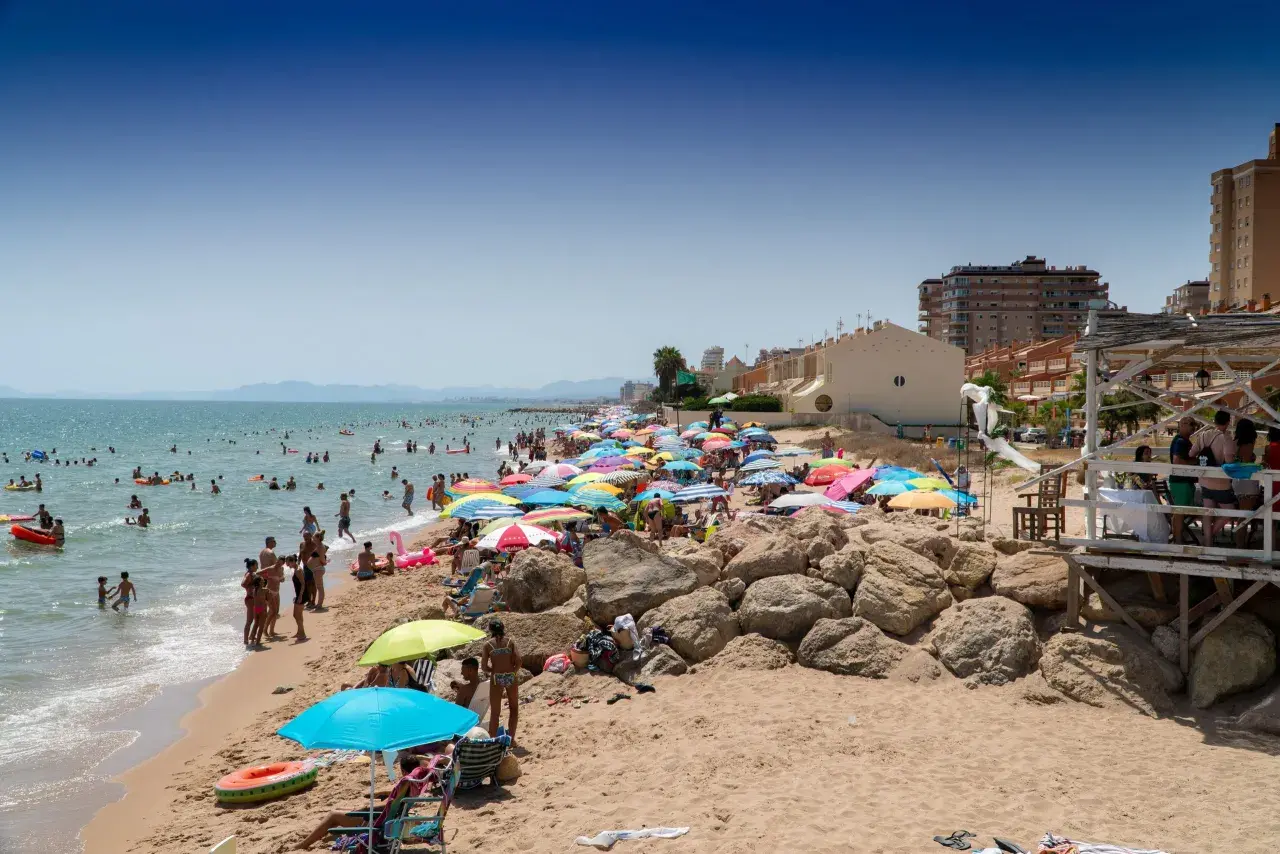 Día soleado en la playa Tavernes de la Valldigna, con mucha gente disfrutando del mar y la arena bajo coloridas sombrillas.