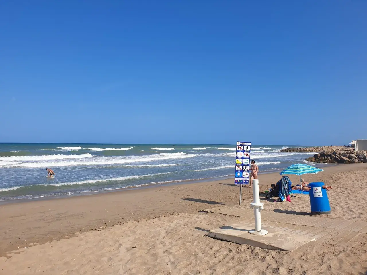 Un día soleado en la playa el Marenyet. Olas rompen en la orilla mientras una persona se baña. Hay un cartel de normas y una sombrilla.