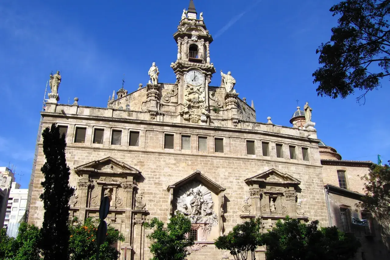 La **iglesia de los Santos Juanes**, con su imponente fachada de piedra, torre con reloj y estatuas, se alza majestuosa bajo un cielo azul intenso.