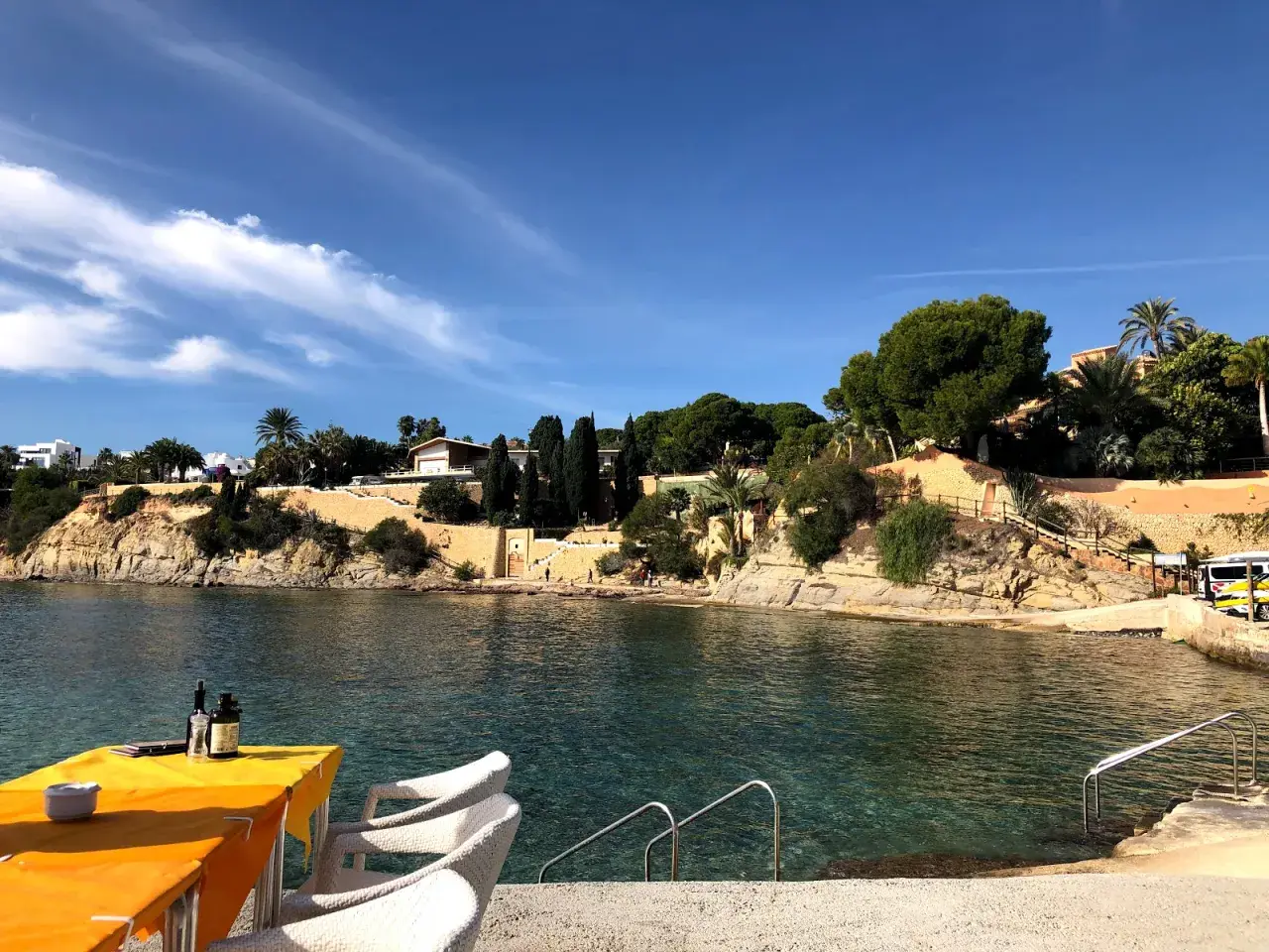 Mesa con mantel naranja y sillas blancas junto al mar cristalino en cala les Bassetes. Casas y vegetación en la ladera.