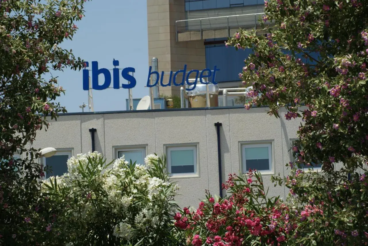 El cartel azul "ibis budget" destaca sobre el edificio del hotel Ibis Budget Alicante, rodeado de exuberante vegetación y flores rosadas y blancas.
