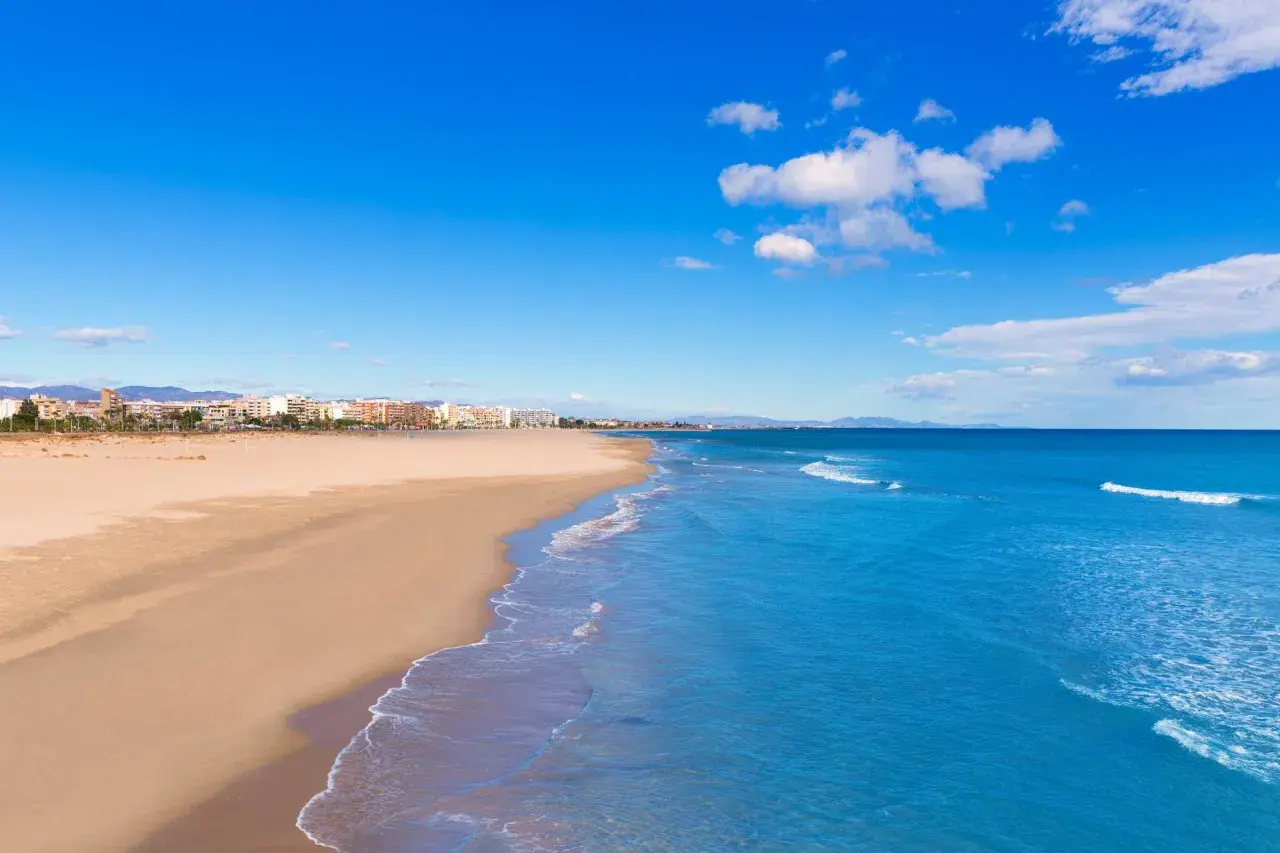Amplia playa de arena dorada en Sagunto, con el mar Mediterráneo azul y edificios de la costa al fondo bajo un cielo despejado.