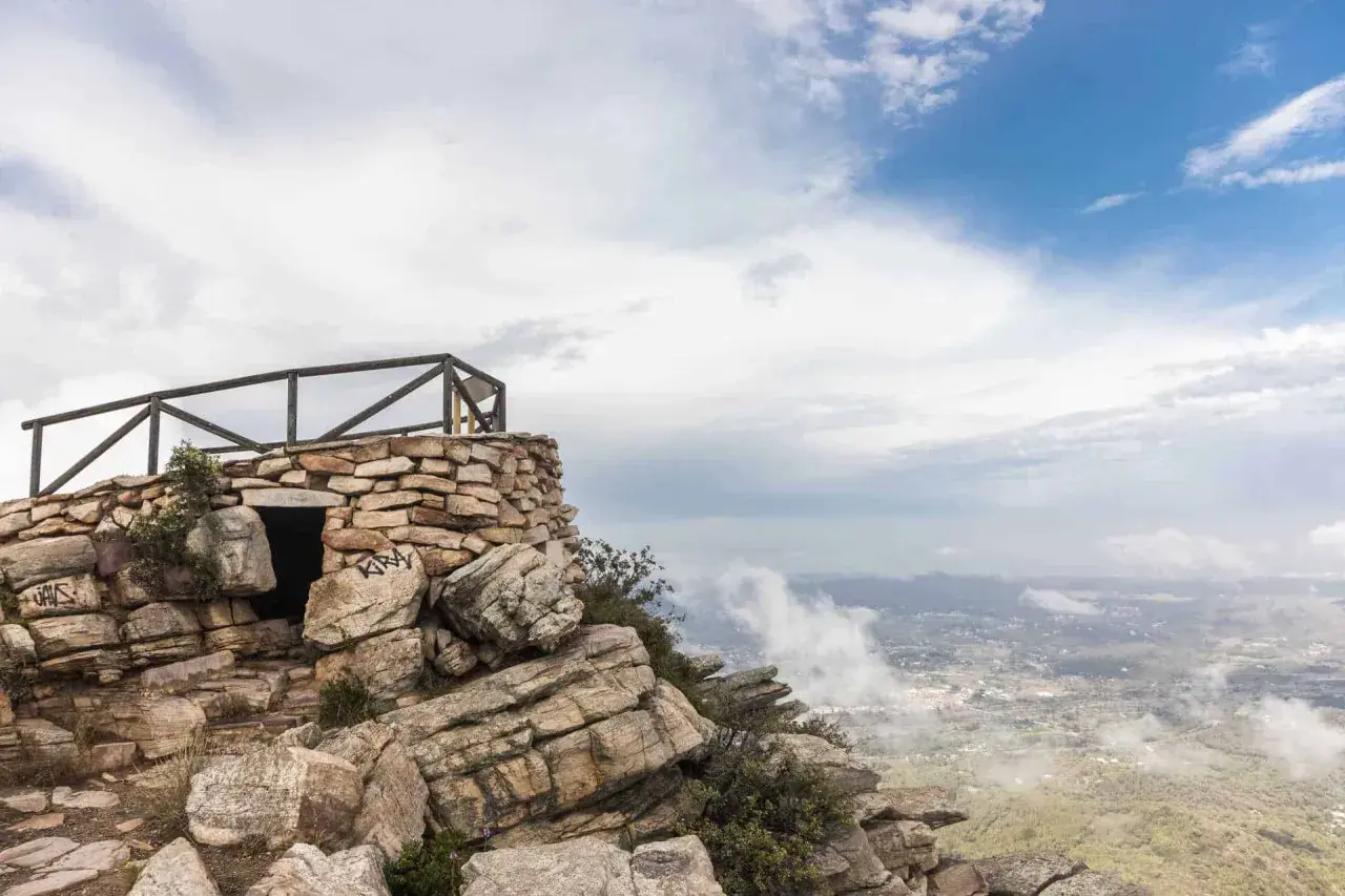 Pequeña construcción de piedra en la cima de una montaña, con barandilla de madera y vistas al valle. El garbí sopla suavemente.