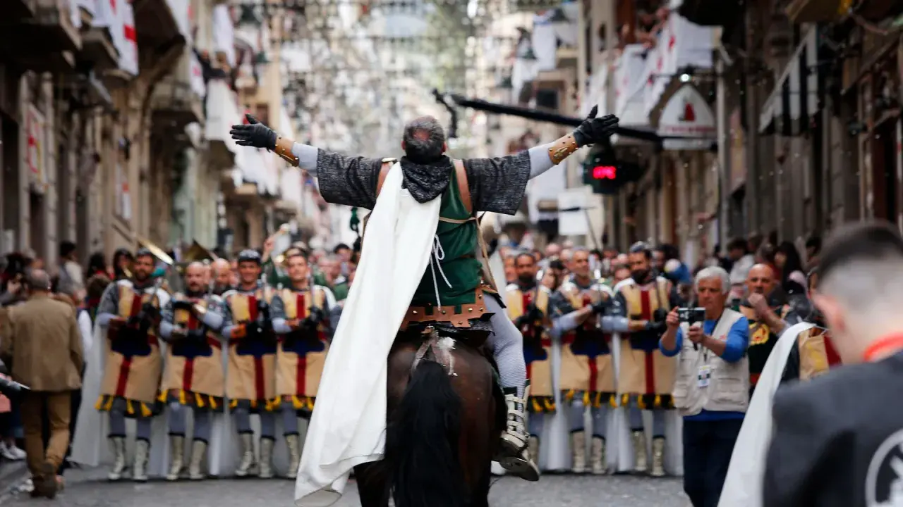 Un jinete con capa blanca lidera un desfile de moros y cristianos, con gente observando y banderas en las calles.