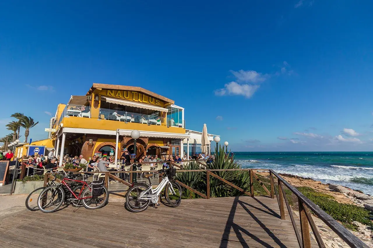 Restaurante Nautilus en la playa Punta Prima, con gente disfrutando en terrazas y bicicletas aparcadas.