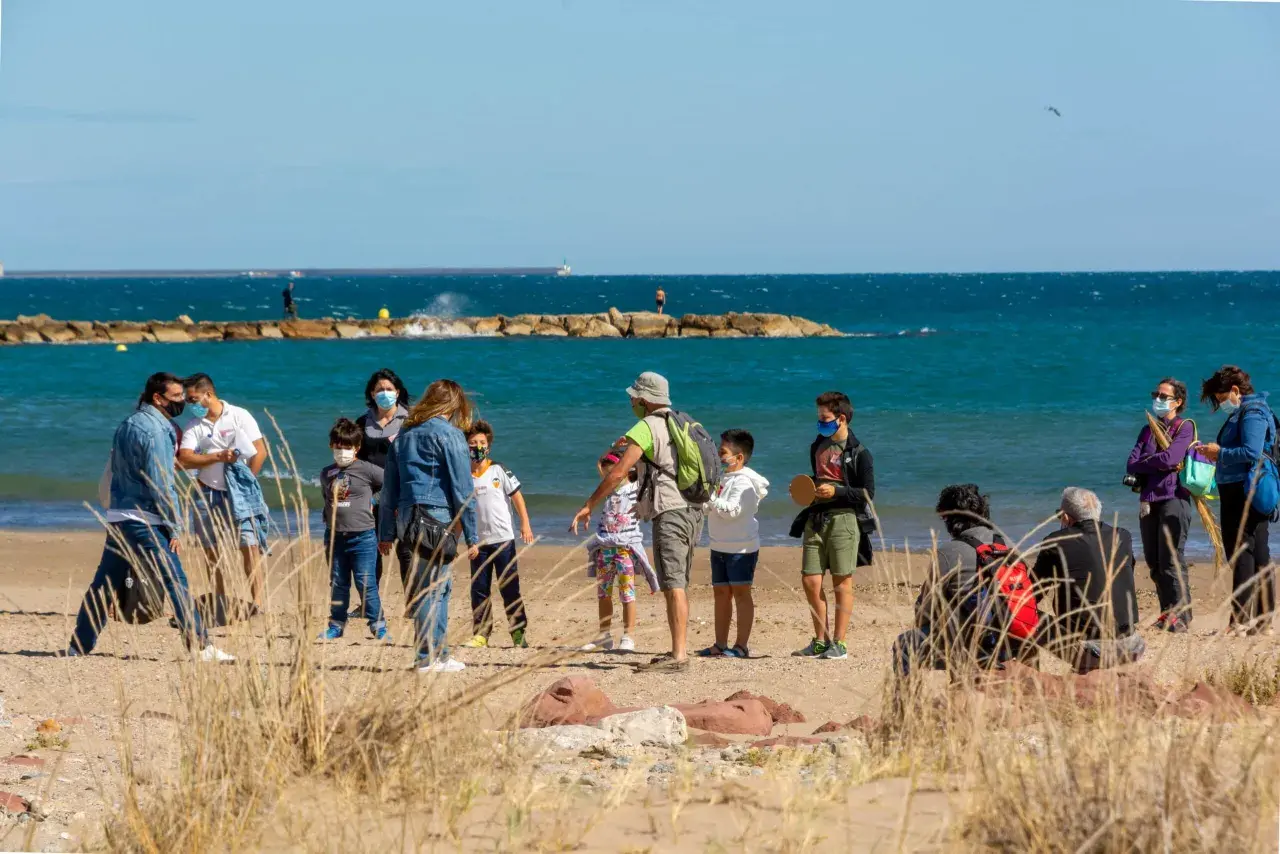 Familias disfrutan de un día soleado en la playa del Puig, con niños jugando y adultos conversando cerca del mar.