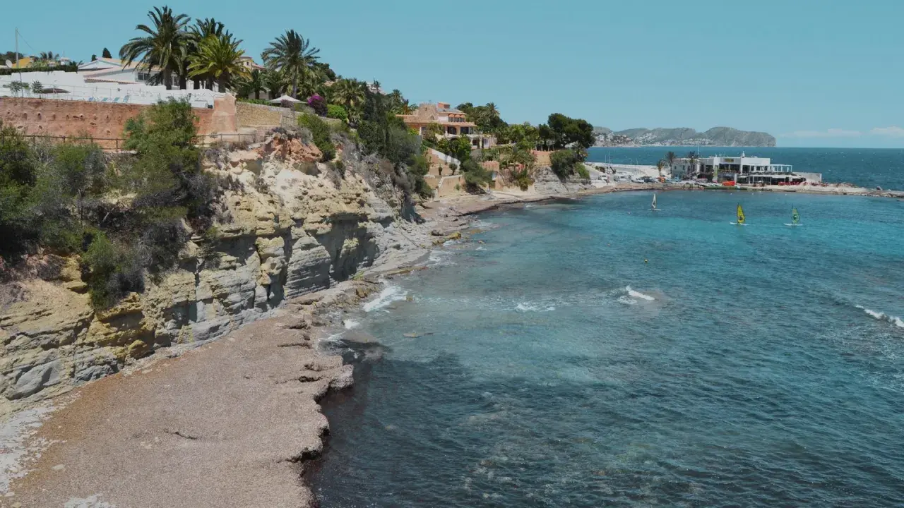 Acantilados y calas rocosas con aguas turquesas, perfectas para descubrir las playas cerca de Calpe. Windsurfistas disfrutan del mar.
