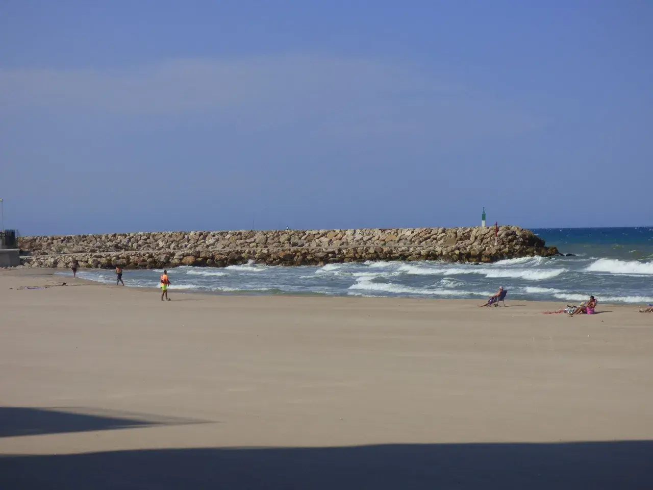 Una playa tranquila con una rompeolas de rocas. Gente relajándose en sillas de playa y paseando. El mar azul y el cielo despejado crean una escena de verano perfecta.