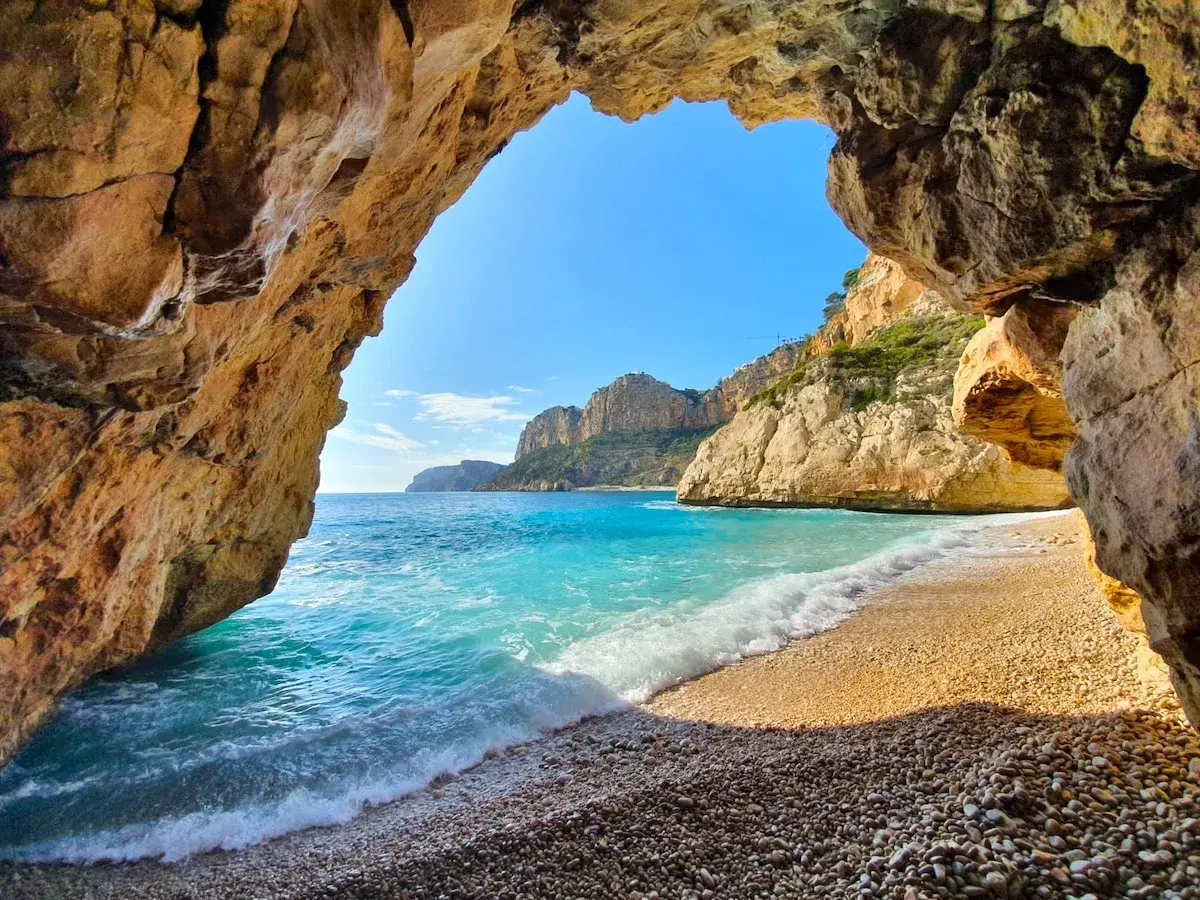 Vista desde una cueva hacia la cala Testos en Benitachell. Aguas turquesas, arena de guijarros y acantilados dorados bajo un cielo azul.