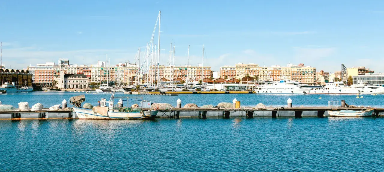 Barcos de pesca y yates de lujo atracados en la marina de Valencia, con edificios coloridos de fondo bajo un cielo azul.