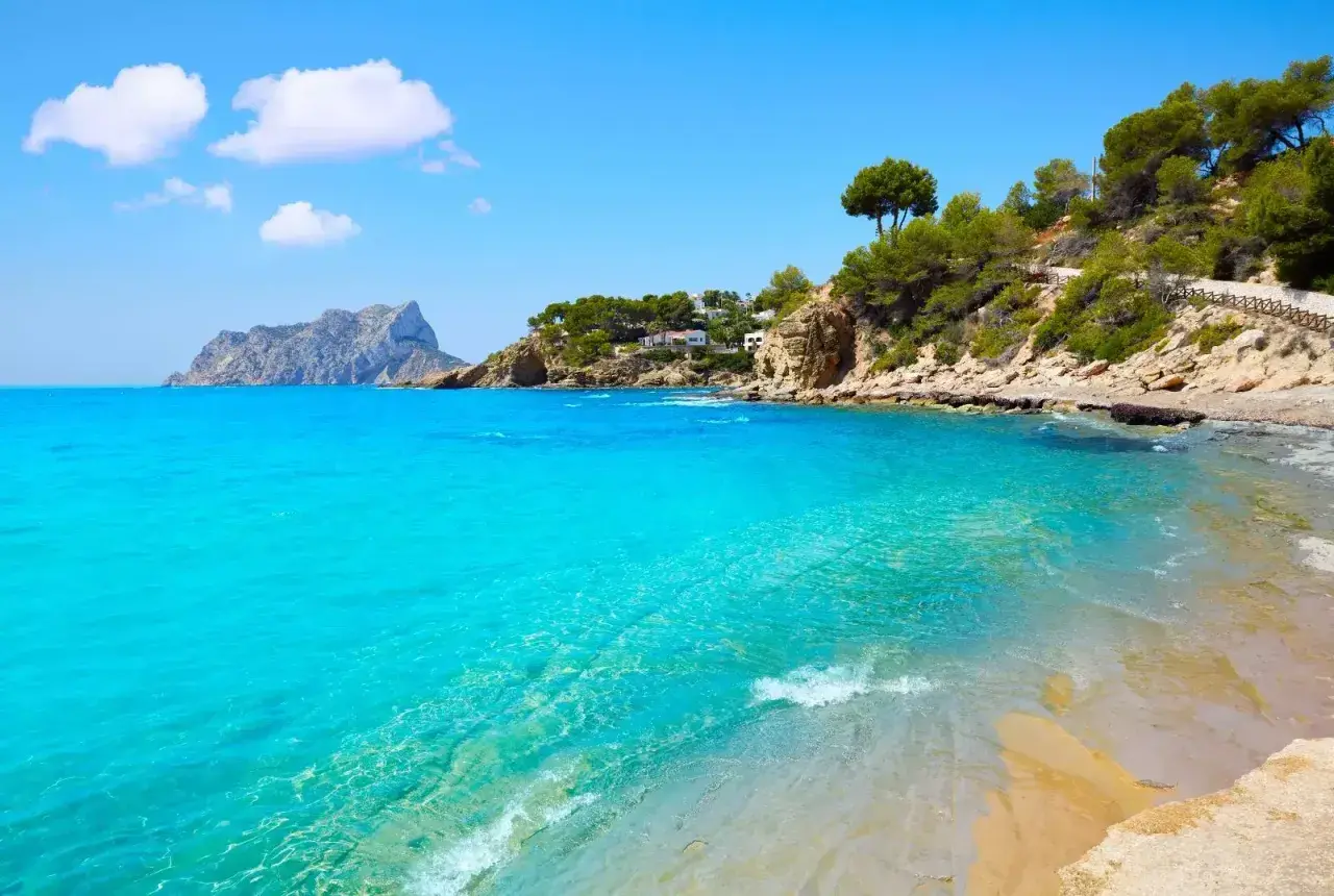 Aguas turquesas y arena dorada en la cala Pinets, con rocas y pinos coronando la costa. Un paraíso mediterráneo.