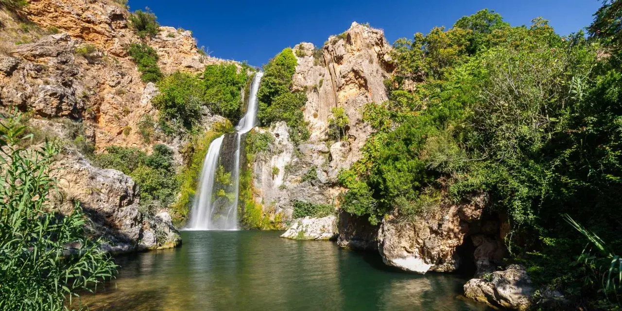 El Salto de Chella, una cascada imponente rodeada de rocas y vegetación exuberante, cae en una poza de agua verde.