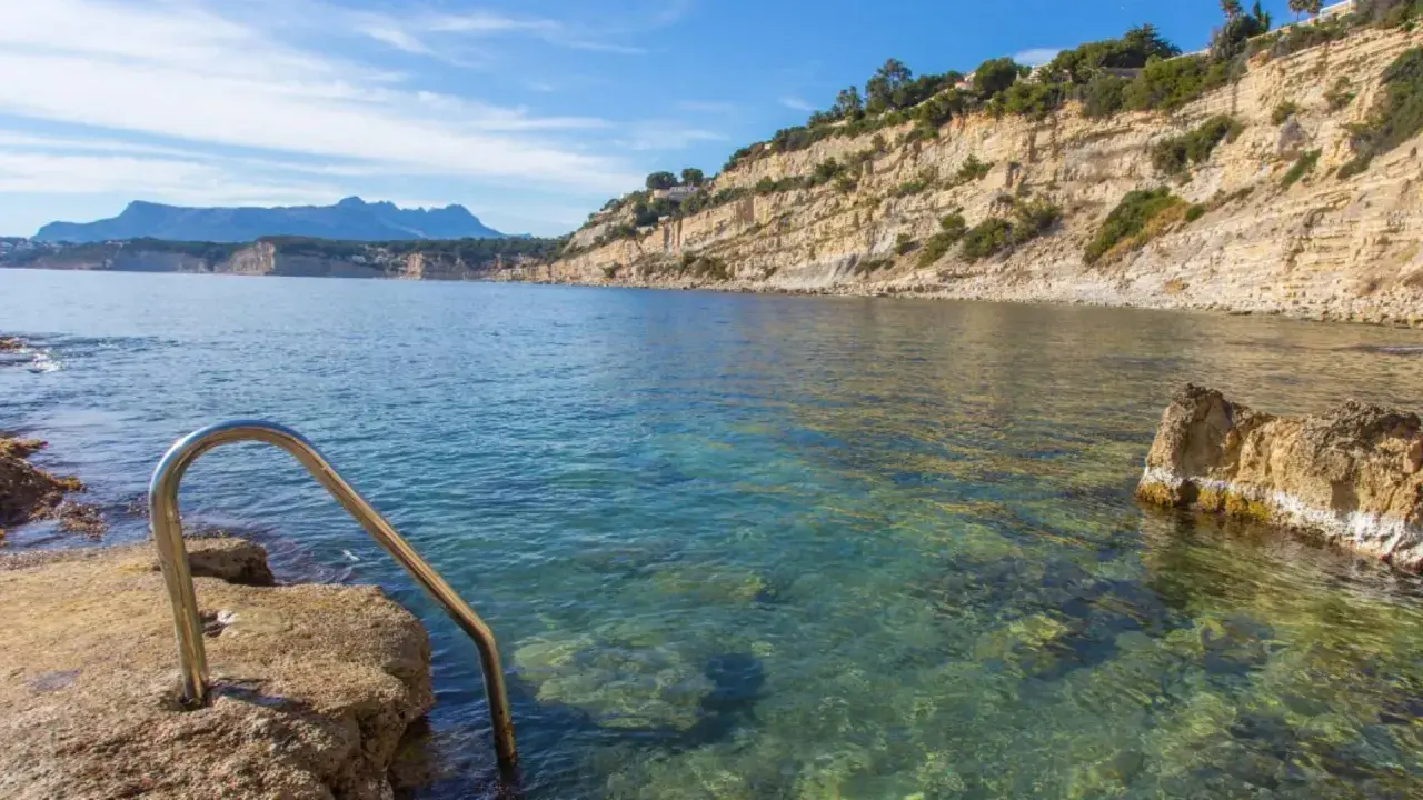 Escalera metálica para acceder a las aguas cristalinas de cala Punta Estrella, con acantilados y montañas al fondo.