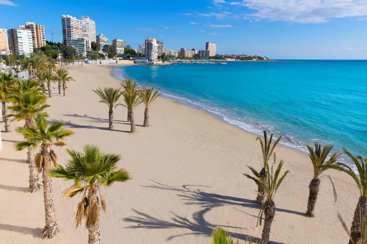 La **playa de Altea** se extiende con arena dorada y palmeras. El mar Mediterráneo, de un azul turquesa intenso, baña la costa, mientras edificios modernos se alzan al fondo.