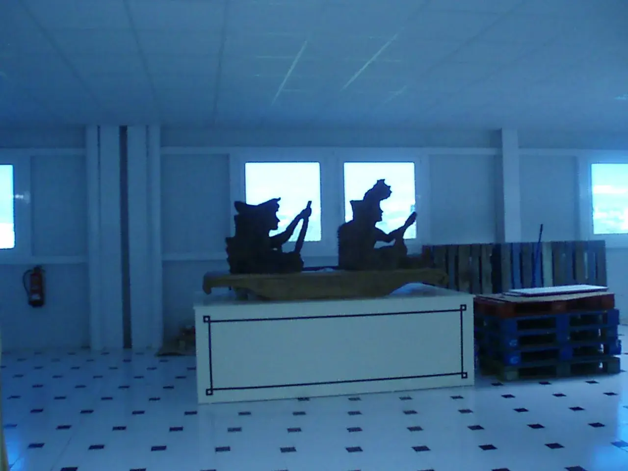 Siluetas de figuras en un interior, quizás una fábrica valor Alicante, con palets y un extintor.