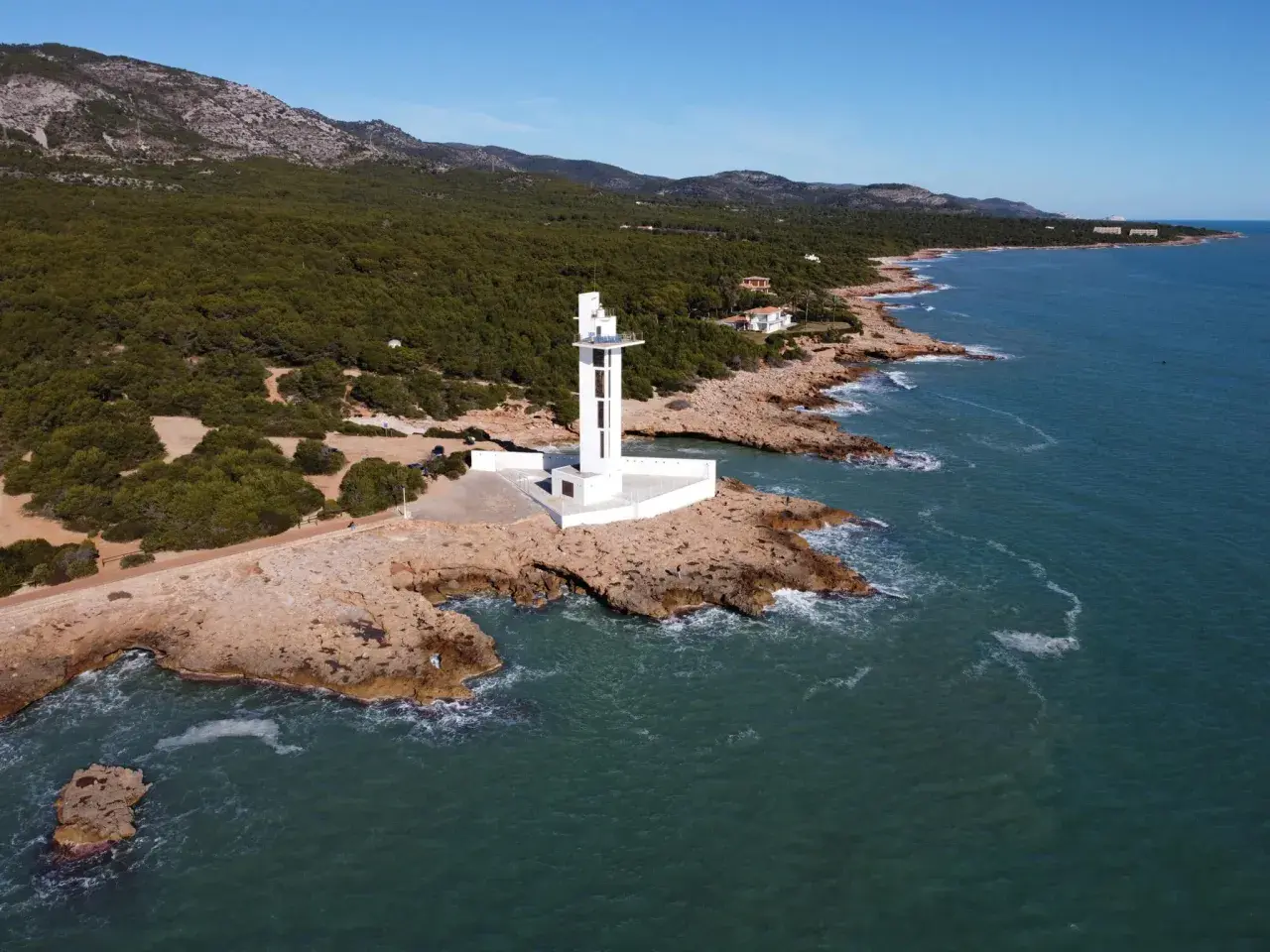 Faro blanco moderno en la costa rocosa de la Sierra de Irta, con el mar azul y montañas verdes al fondo.
