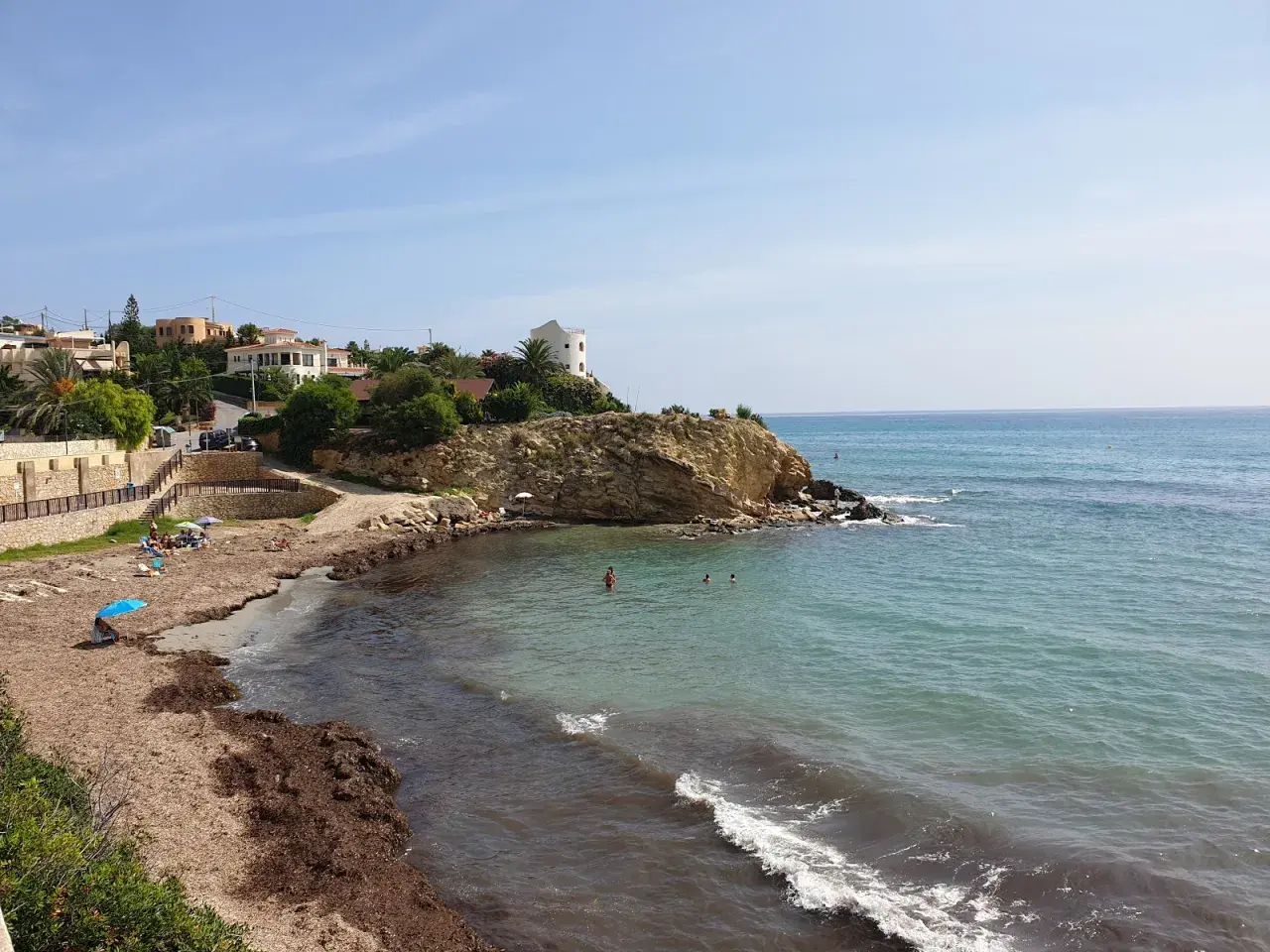 La cala Coveta, con su playa de arena y rocas, invita a disfrutar del mar Mediterráneo. Casas blancas coronan la colina.