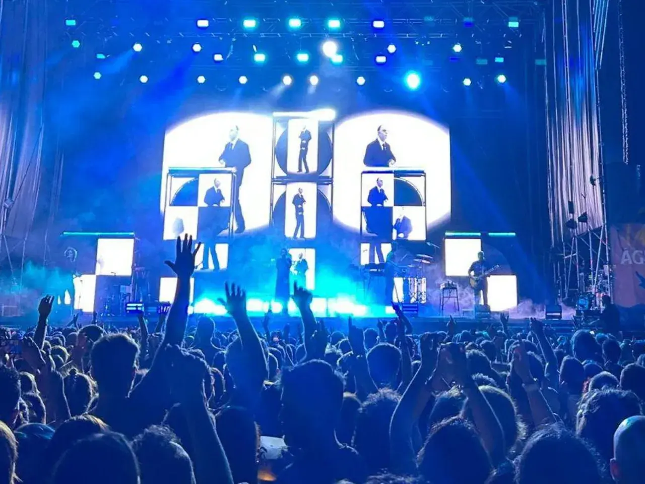 Multitud en concierto de Vetusta Morla en Valencia. Luces azules y pantallas con imágenes de los miembros de la banda crean un ambiente electrizante.