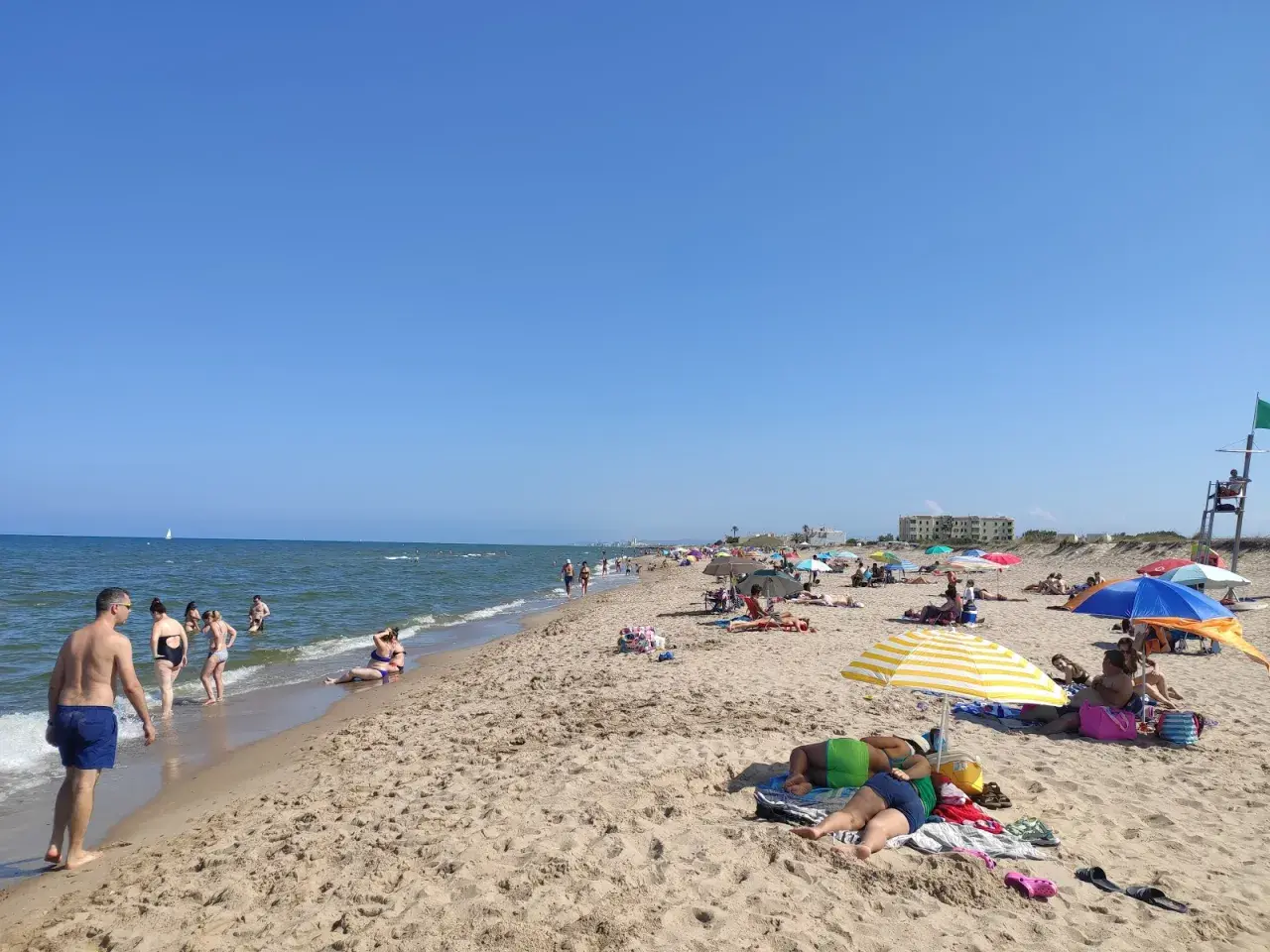 Gente disfrutando del sol y el mar en la playa de la Garrofera. Sombrillas, toallas y bañistas llenan la arena dorada bajo un cielo azul.