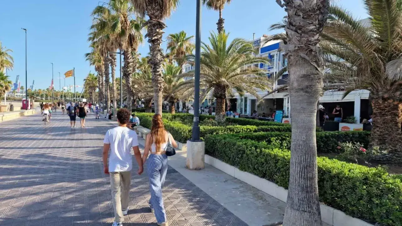 Pareja paseando por el paseo marítimo de Valencia, con palmeras, gente y edificios al fondo.