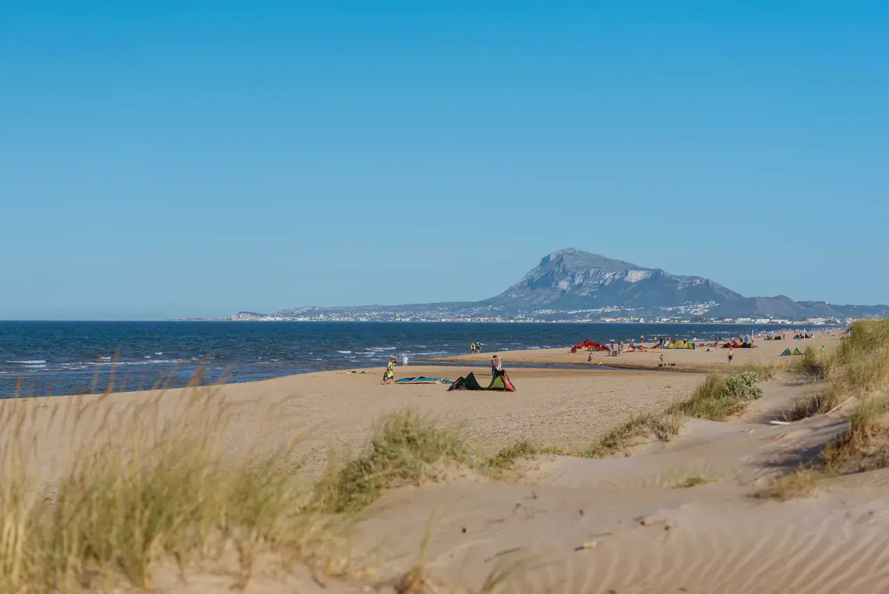 Dunas y mar en la playa de Rabdells, con gente disfrutando del sol y el paisaje montañoso al fondo.