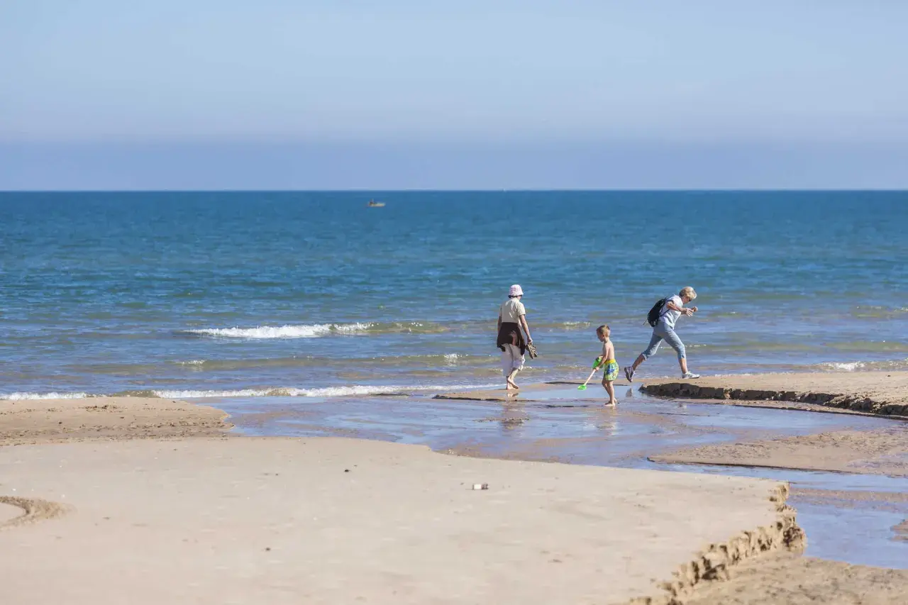 Un niño y dos adultos disfrutan de un día soleado en la playa Pau Pi, jugando cerca de la orilla del mar azul.
