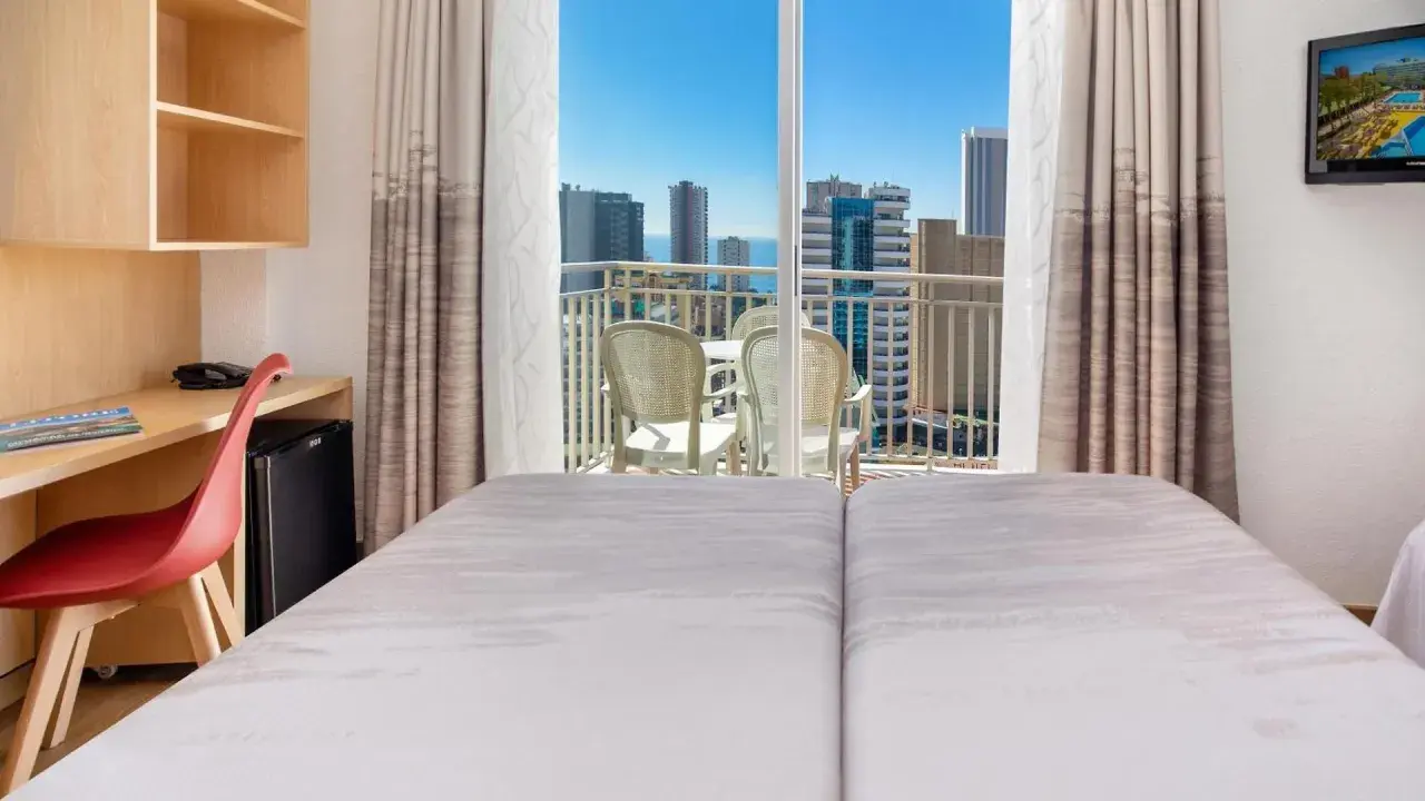 Habitación de hotel pueblo Benidorm con vistas al mar y rascacielos. Balcón con mesa y sillas. Escritorio con silla roja y minibar.